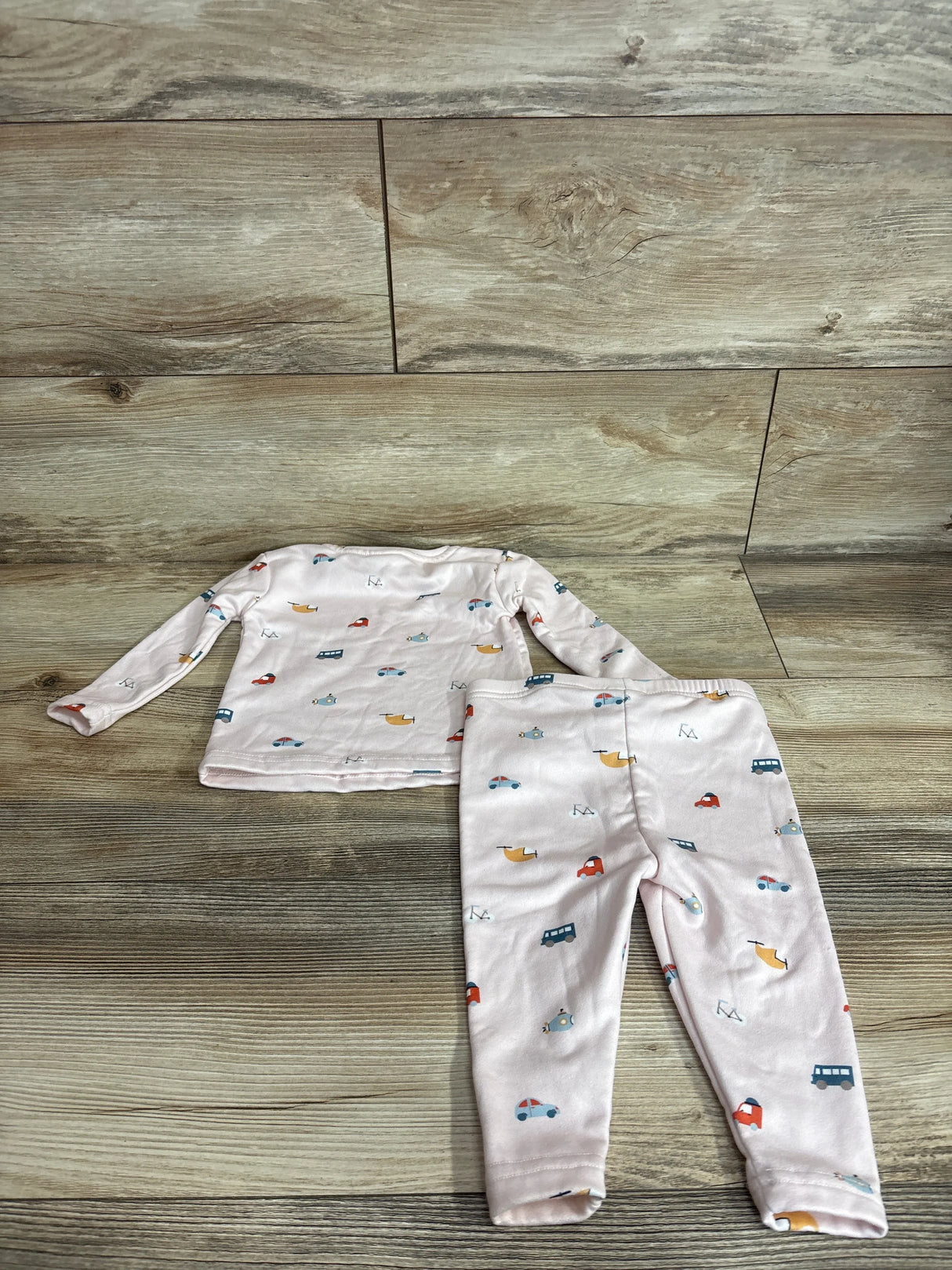 Top-down view of pink 2-piece baby pajama set showcasing long-sleeve top and elastic-waist pants with small multicolored vehicle print on wood background