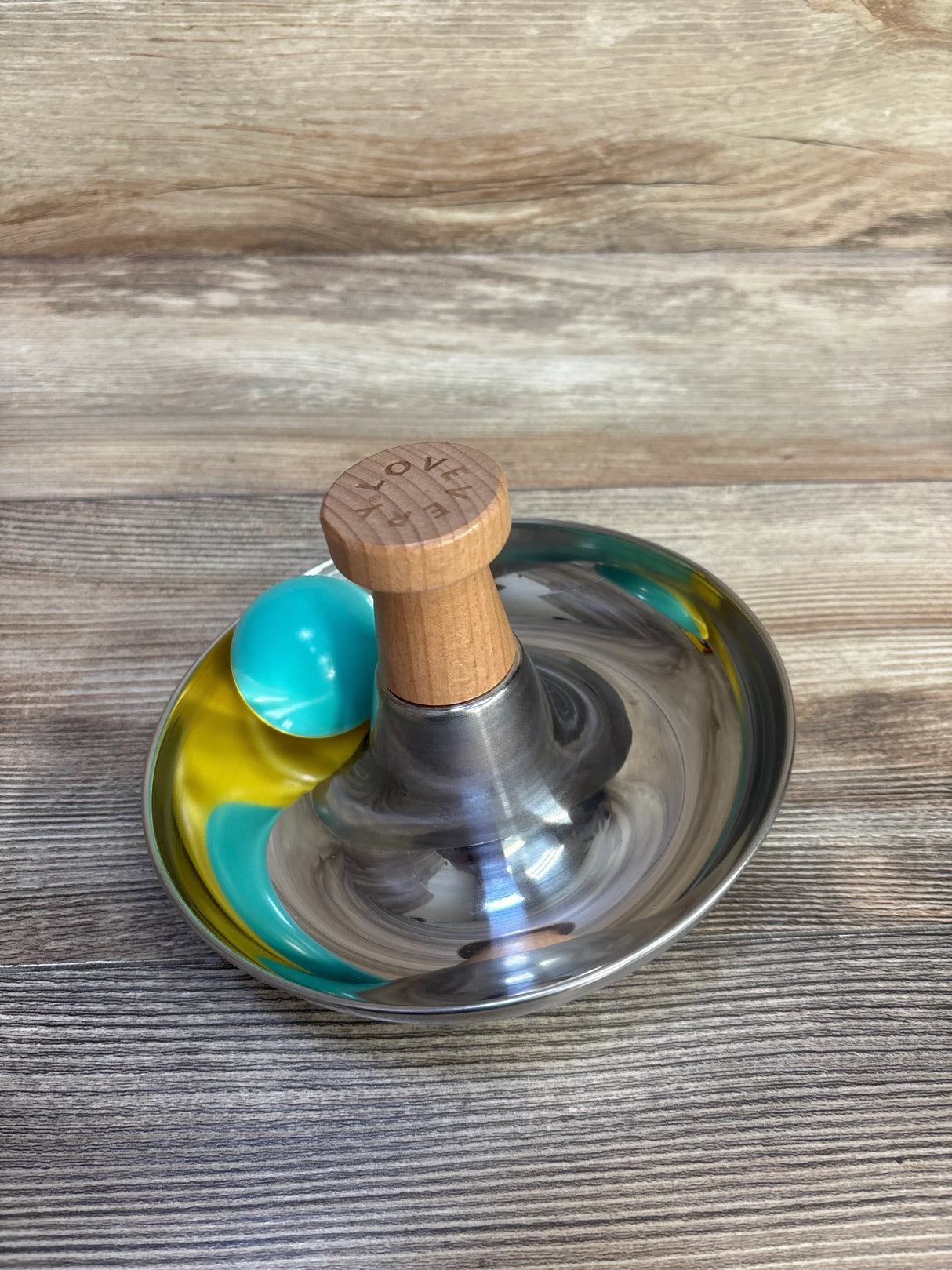 Angled view of Lovevery Stainless Steel Tip and Turn: stainless bowl, wooden LOVE knob, teal-yellow ball on wood background