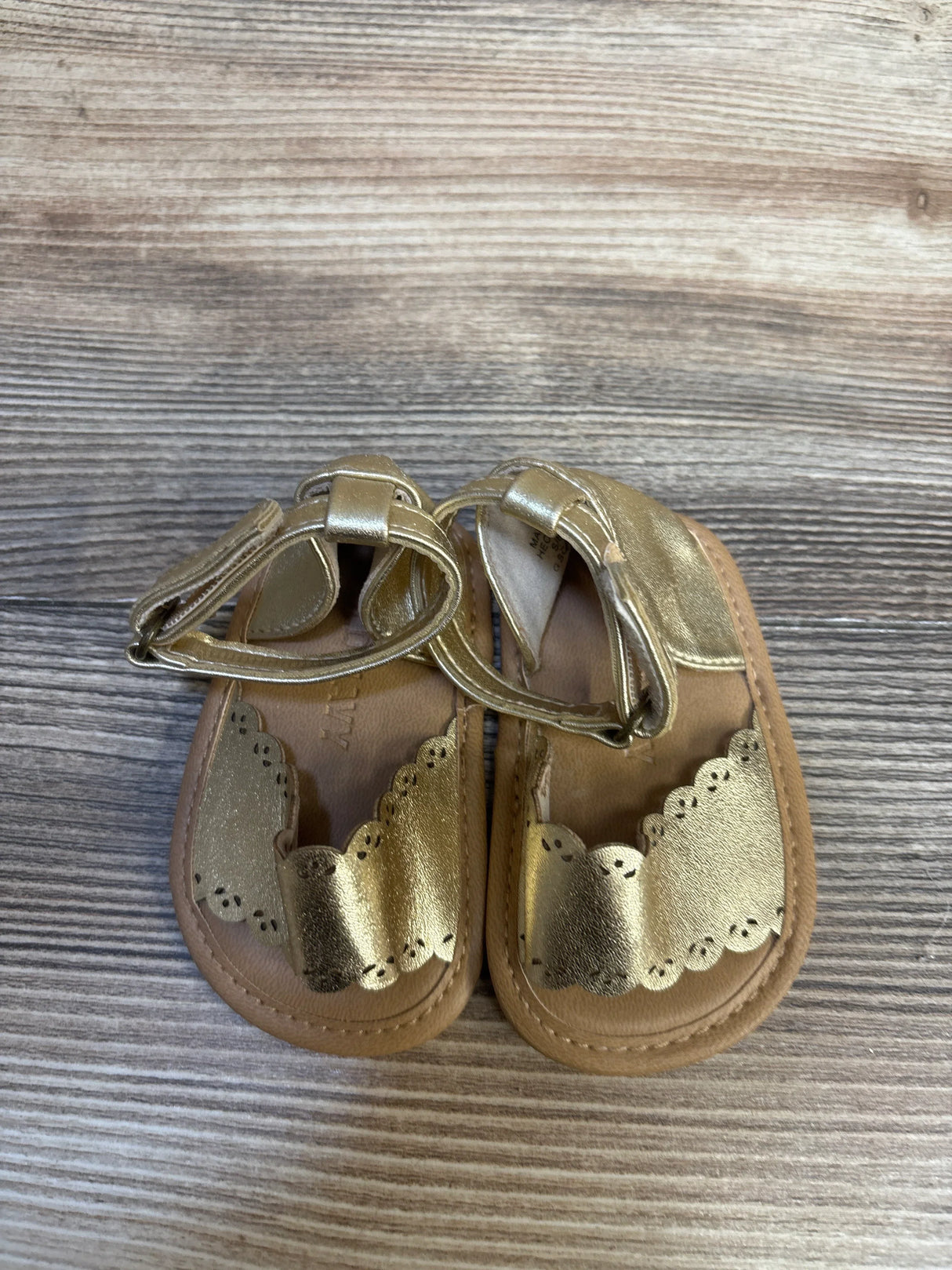 Top-down view of Old Navy metallic faux-leather scallop-trim baby sandals, size 1/2C (0–3M), showing ankle straps on wood background