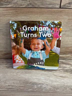 Front view of Lovevery Graham Turns Two board book showcasing the cover photo of a toddler with paint-covered hands with a wooden background