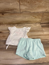 Top-down view of a two-piece toddler shirt and shorts set with a white ruffled-sleeve shirt on a wood floor