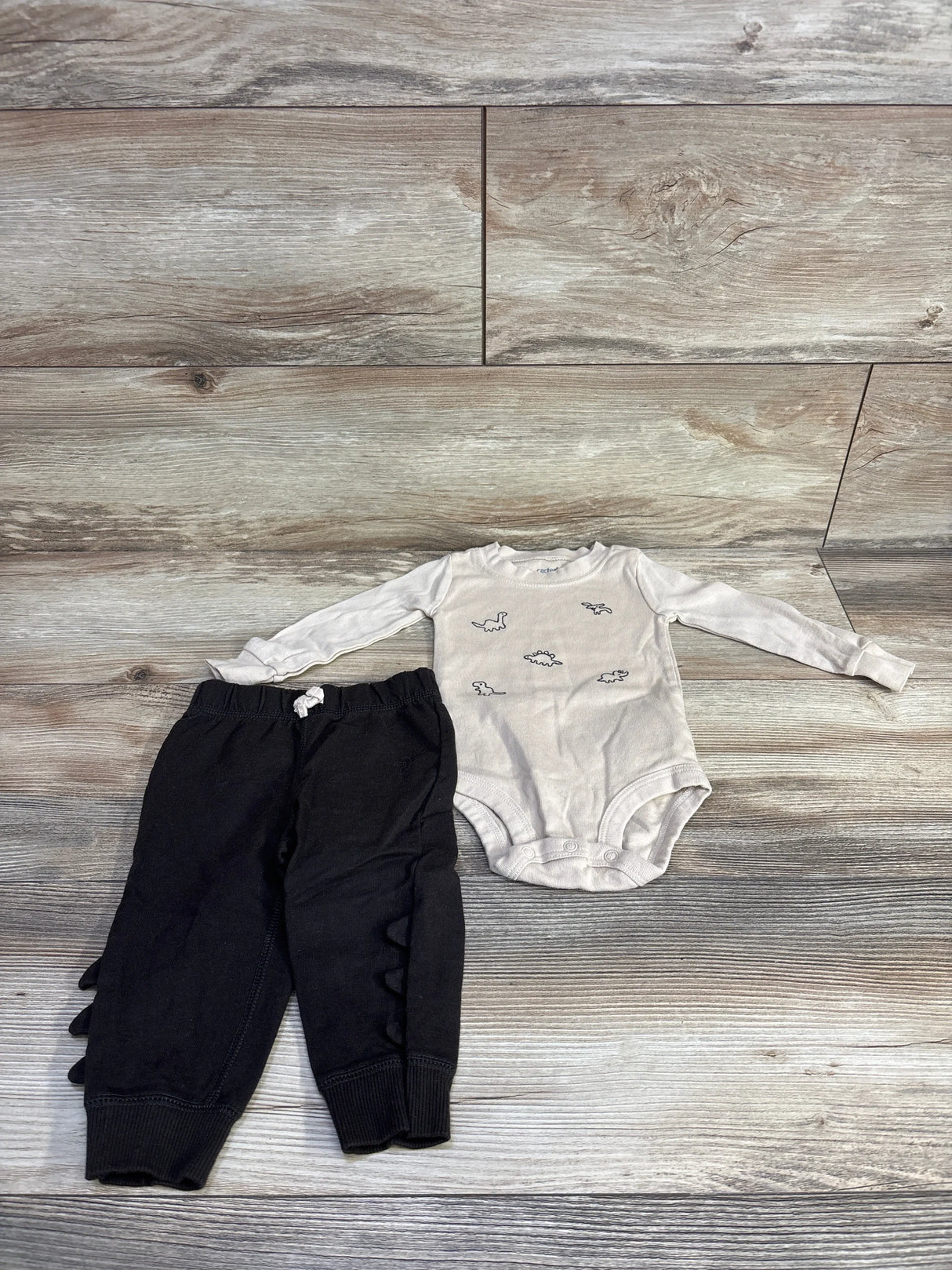 Flat lay of beige long-sleeve dinosaur bodysuit and black pants showing dinosaur prints and ruffle spikes on light wood