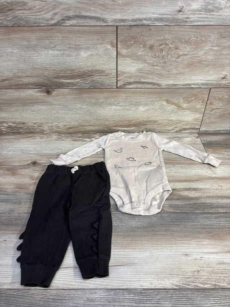 Flat lay of beige long-sleeve dinosaur bodysuit and black pants showing dinosaur prints and ruffle spikes on light wood