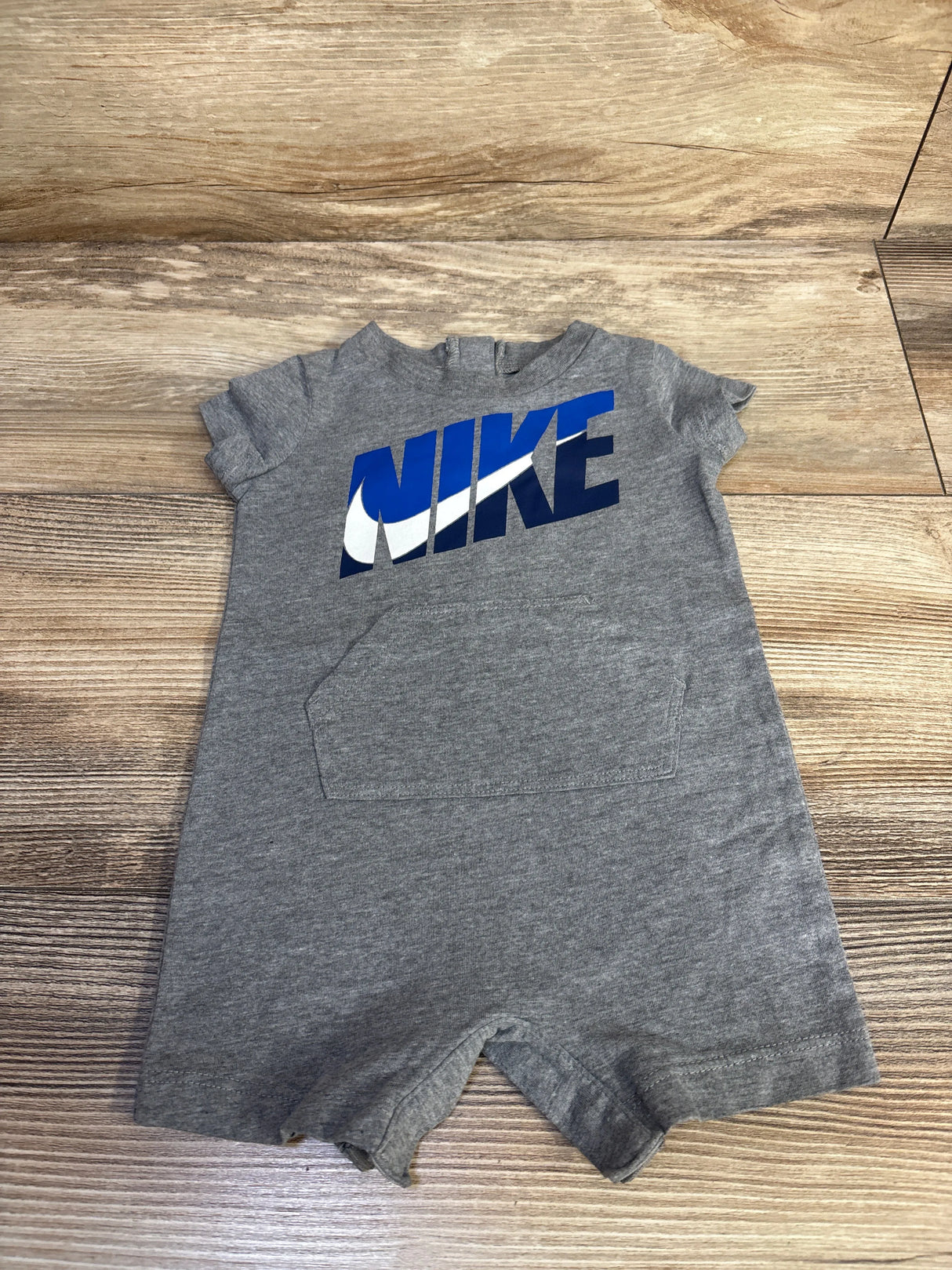 Top-down view of grey Nike shortie romper size 3M showing blue-and-white NIKE logo and front kangaroo pocket on wood floor