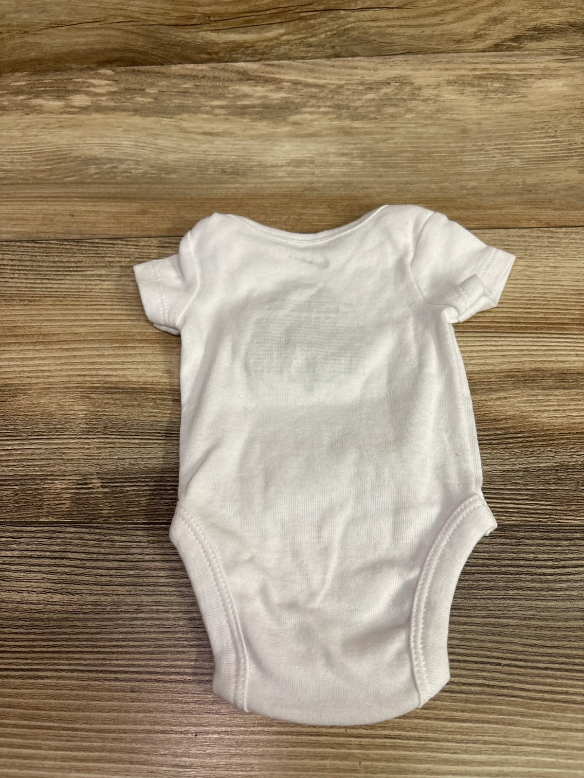 Front view of Nike white newborn bodysuit showcasing short sleeves and rounded neckline with a wood-grain background