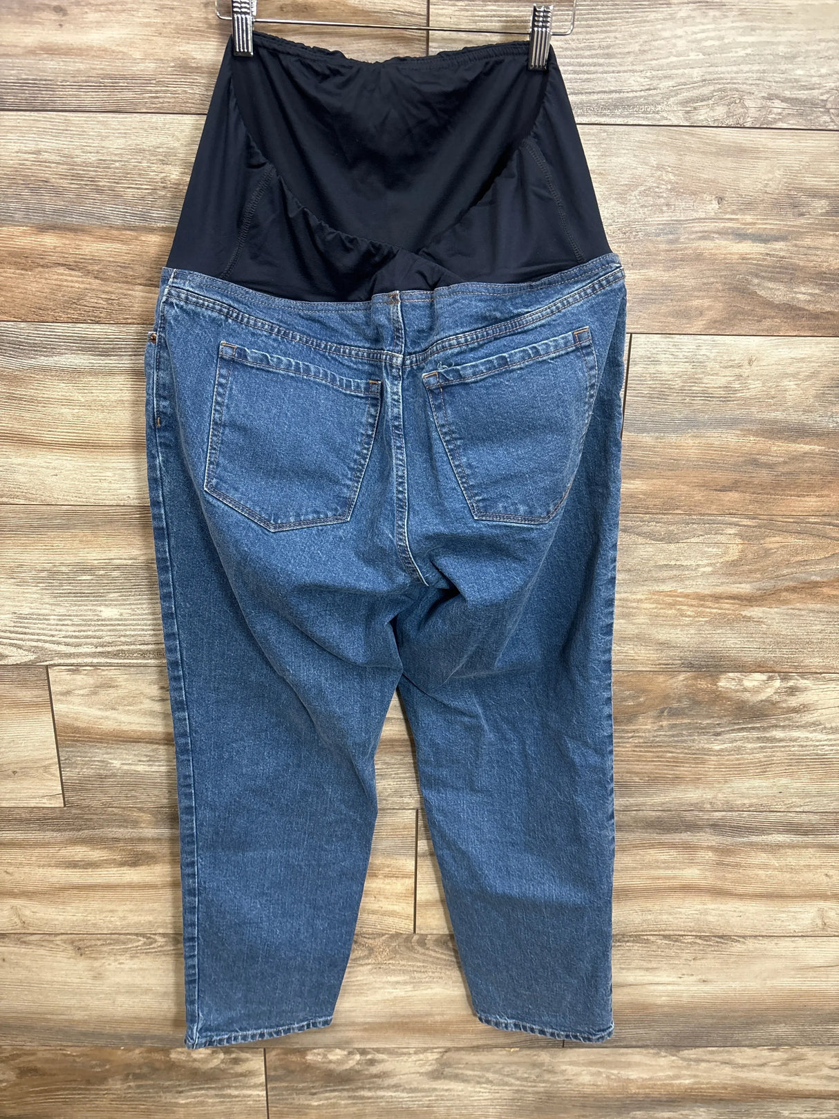 Back of Isabel Maternity 90s straight jeans showing black over-belly panel and blue denim on hanger against wood background