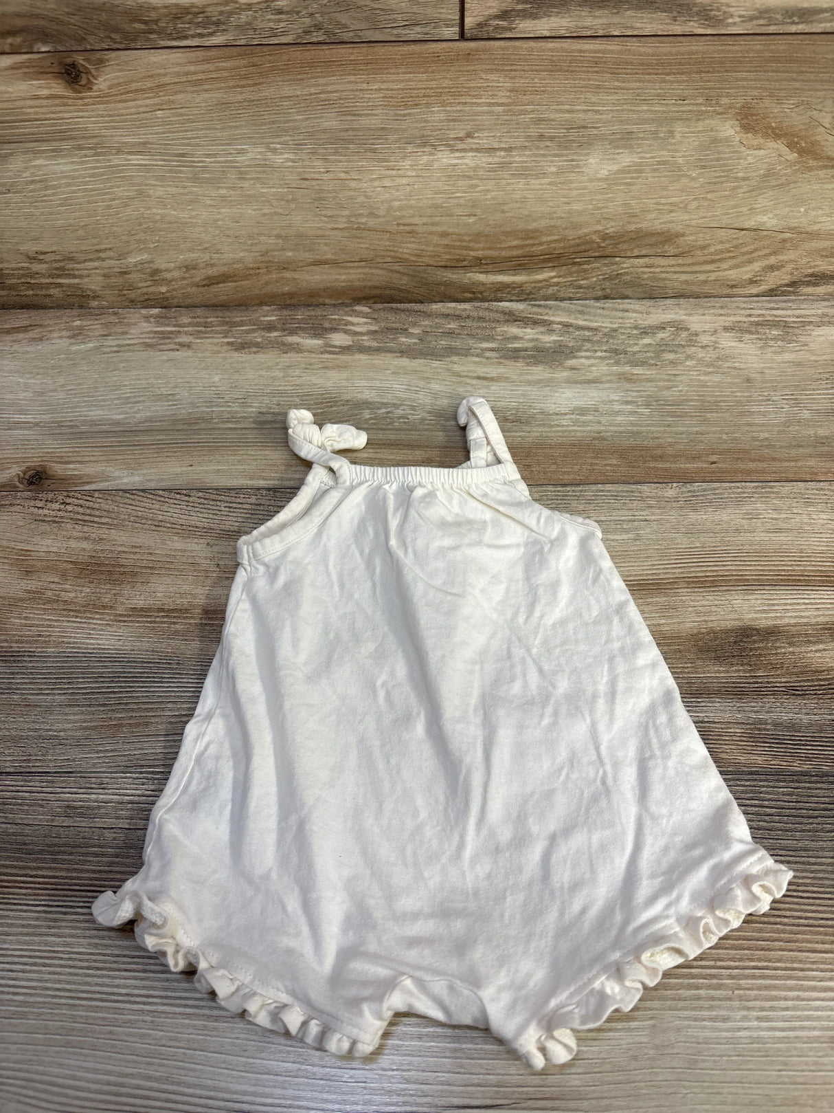 Flat lay of ivory baby tank shortie romper showing knotted shoulder straps and ruffle hem at legs on wood background
