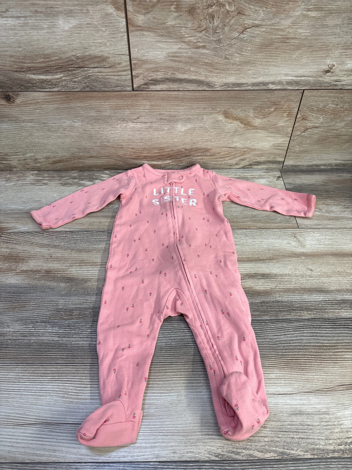 Top-down view of pink footed baby sleeper with front zipper, small floral print and 'LITTLE SISTER' text on wood background