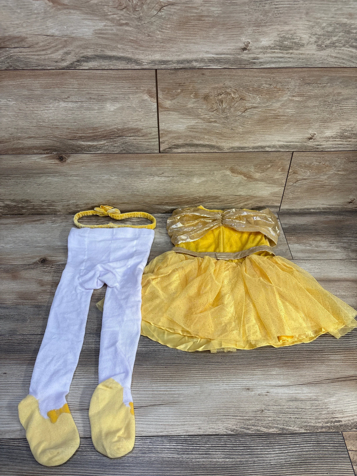 Flat-lay of toddler Belle three-piece costume: glitter gold bow bodice, yellow tulle skirt, white tights and headband on wood floor