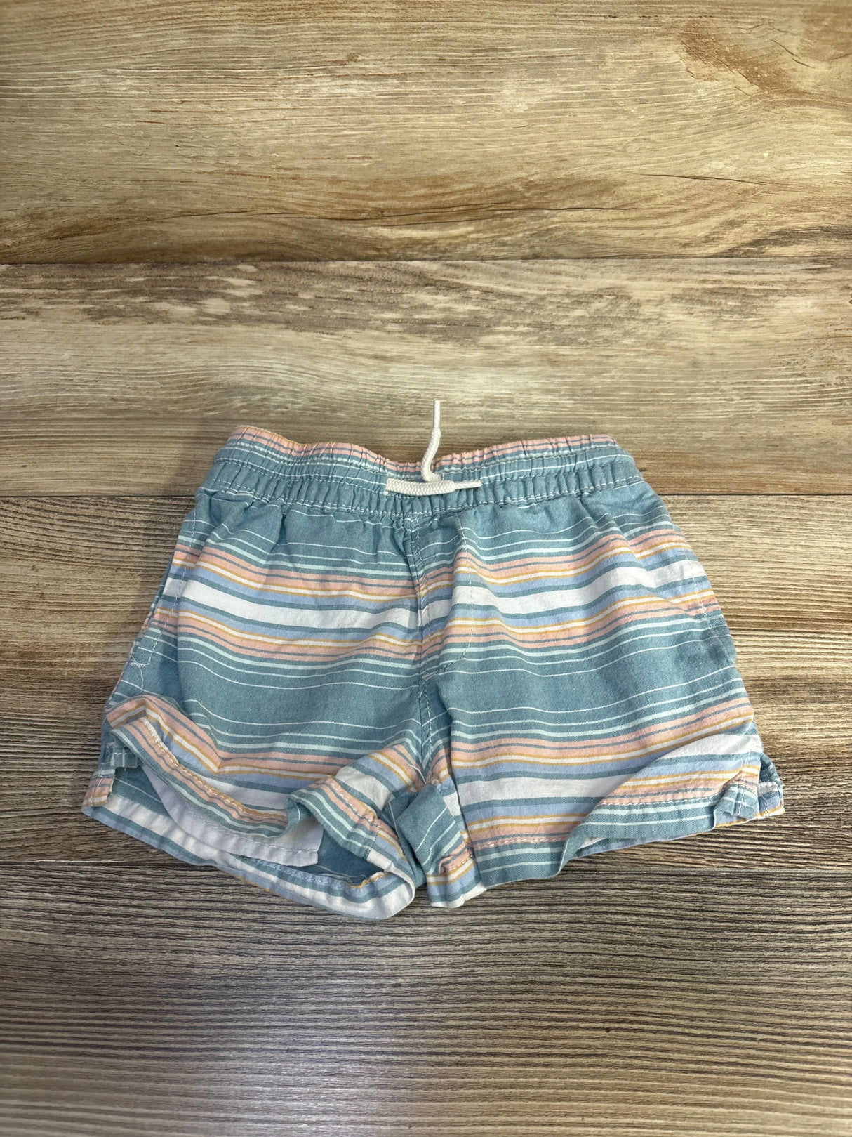 Front view of OshKosh boys' blue striped cotton shorts with elastic waistband and white drawstring on wood background