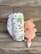 Front view of plush baby blanket rolled beside fox plush showing woodland animal print and white sherpa back on wood backdrop