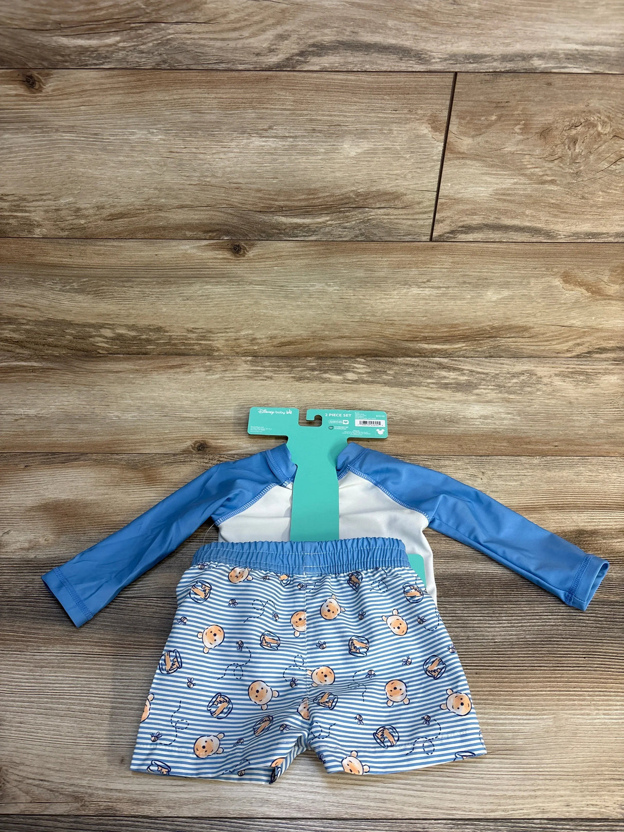 Flat lay of light blue Disney Baby 2-piece Winnie the Pooh rash guard set showing long-sleeve top and striped shorts on wood background