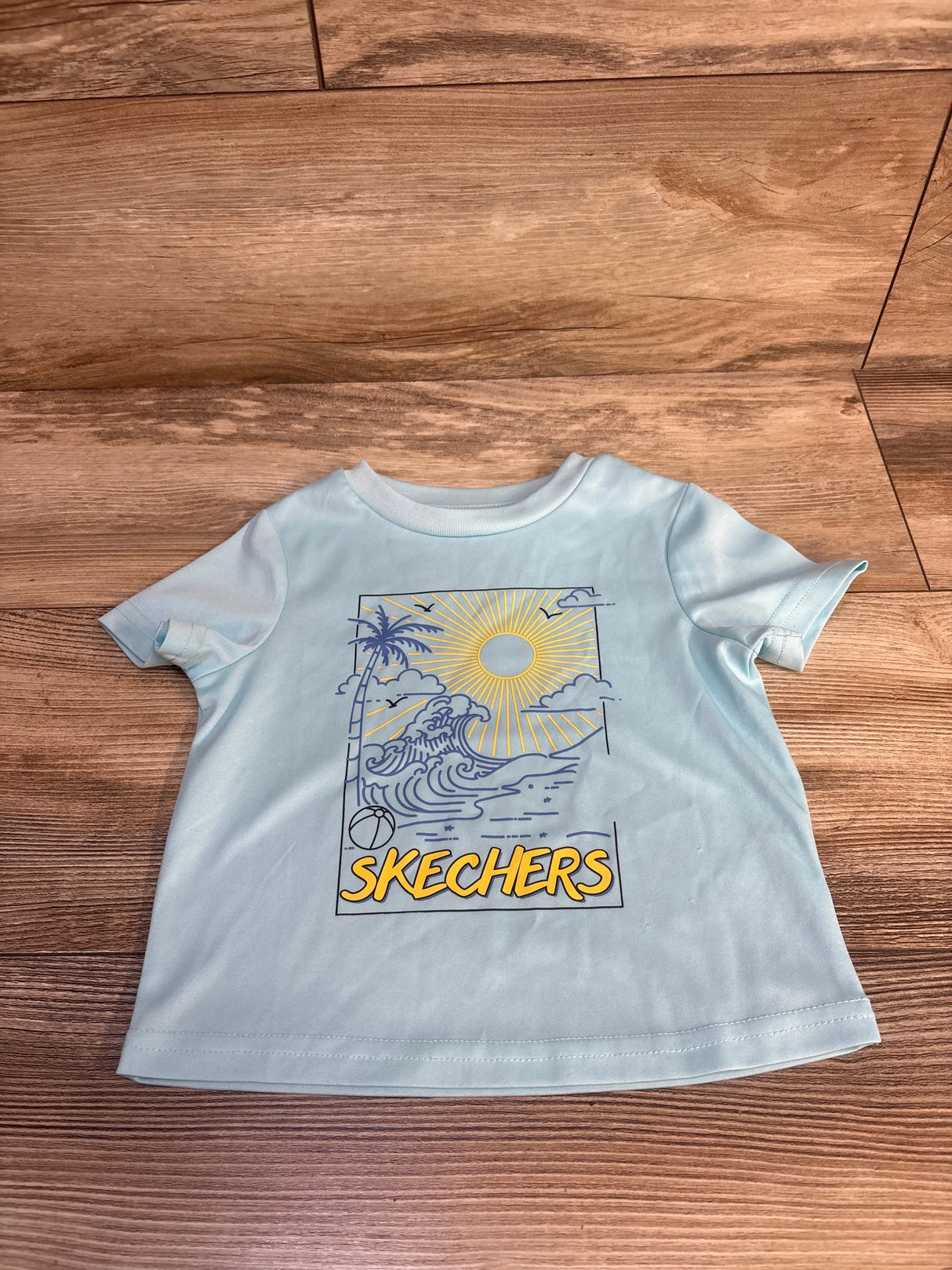 Light blue kids Skechers t-shirt with beach graphic, sun, palm tree, waves, and beach ball on wood floor