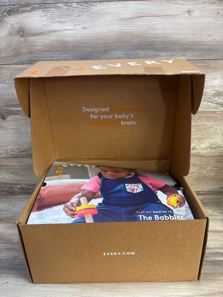 Front view of Lovevery The Babbler play kit box open showing brochure with toddler holding wooden ring toy on wood background