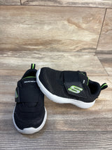 Black Skechers toddler athletic shoes with green accents and Velcro straps on wood floor