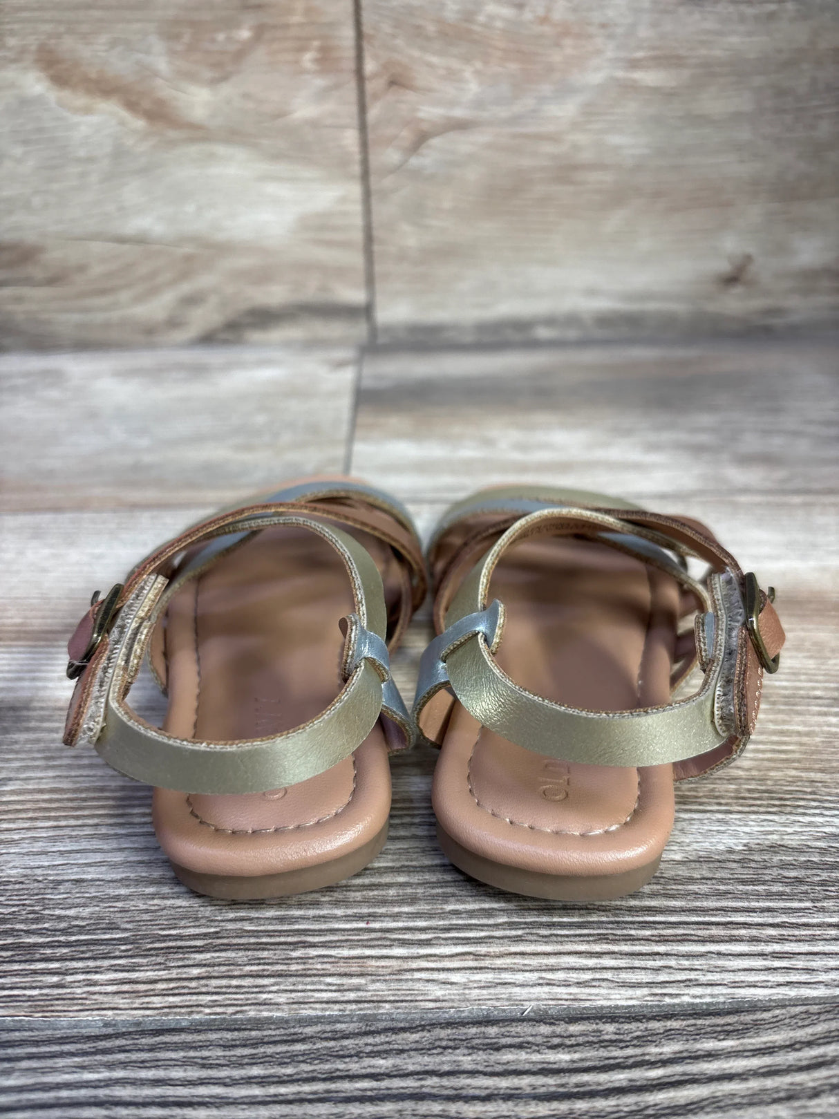 Rear view of toddler metallic gold faux-leather strappy sandals size 7C showing ankle buckle straps and padded tan footbed on wood plank background