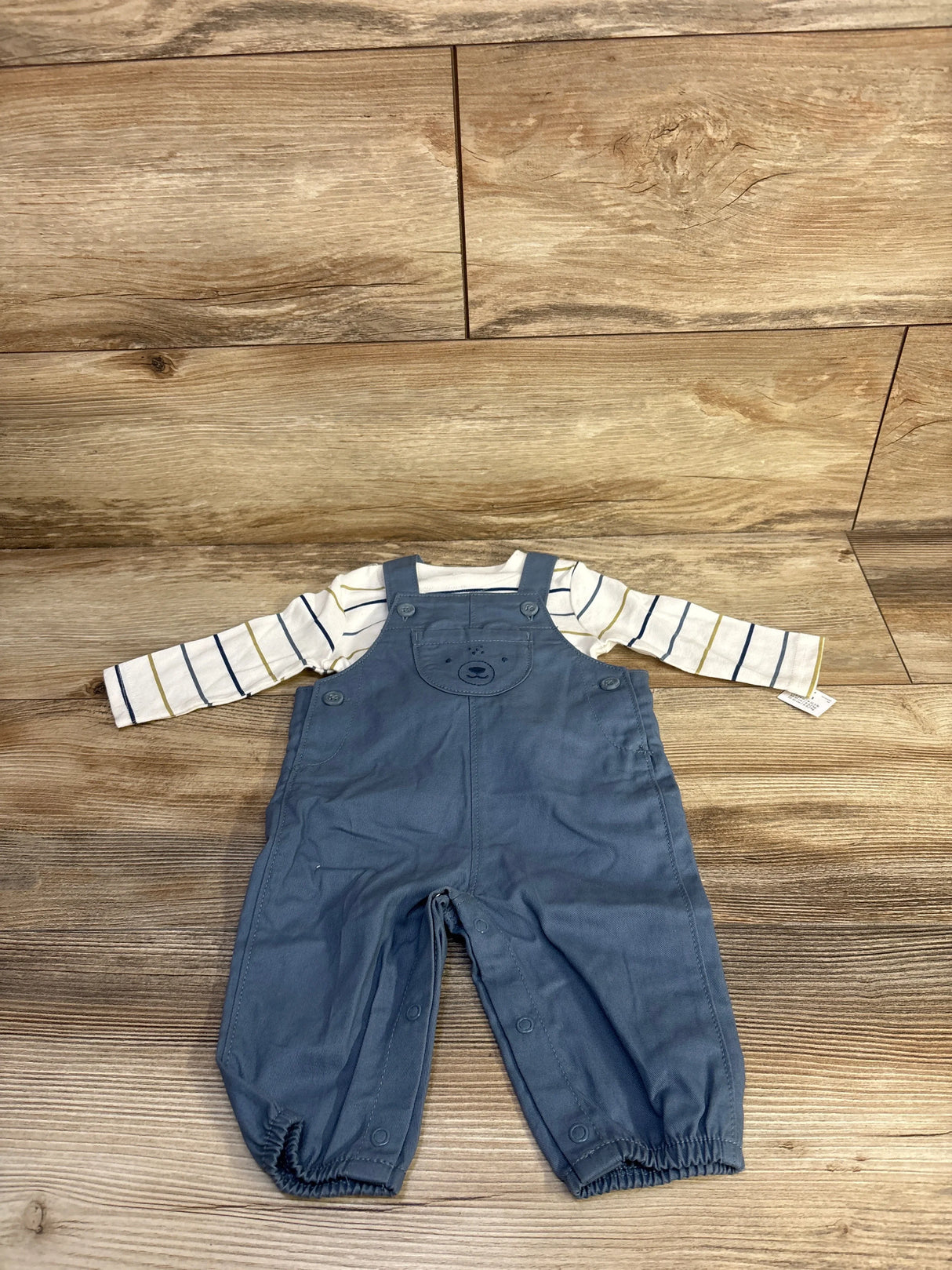 Top-down view of baby 2-piece: white striped long-sleeve shirt and blue overalls with bear bib and leg snaps on wood