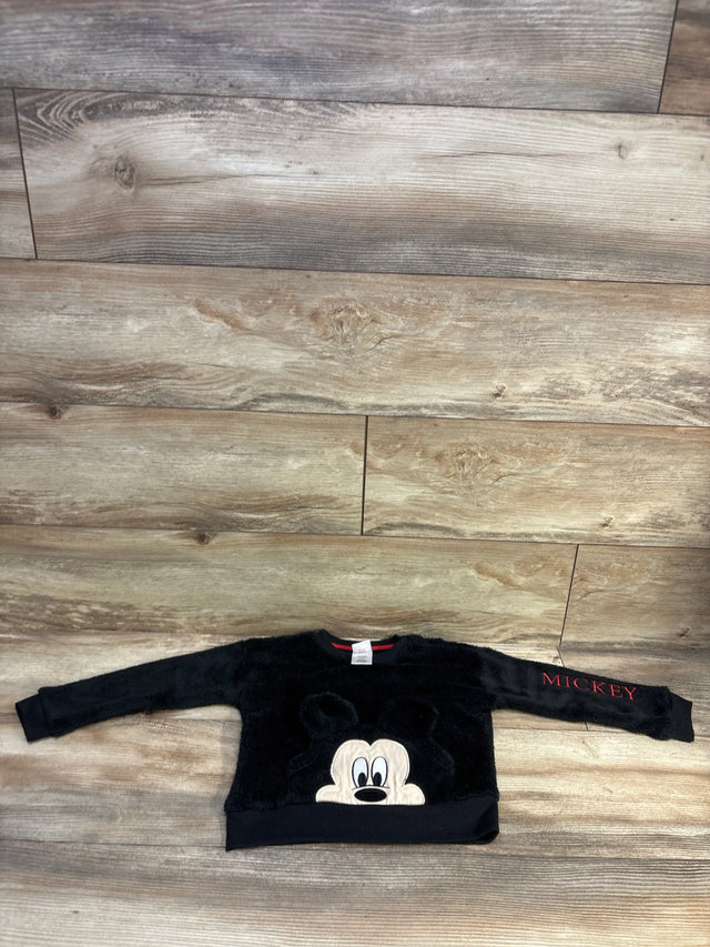 Flat lay of black toddler plush Mickey Mouse pullover sweatshirt showing front Mickey face applique and red MICKEY sleeve text on wood floor
