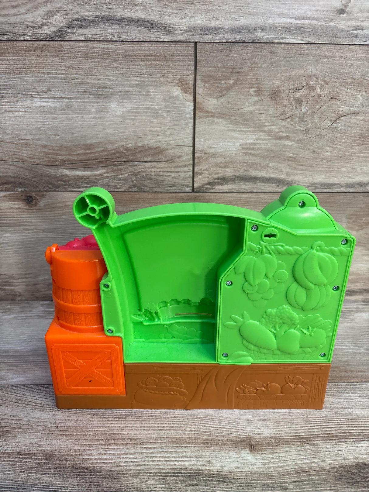 Rear view of Fisher Price Laugh & Learn Fruits & Fun Learning Market showcasing green market interior, orange base, and removable fruit bin on wood plank background