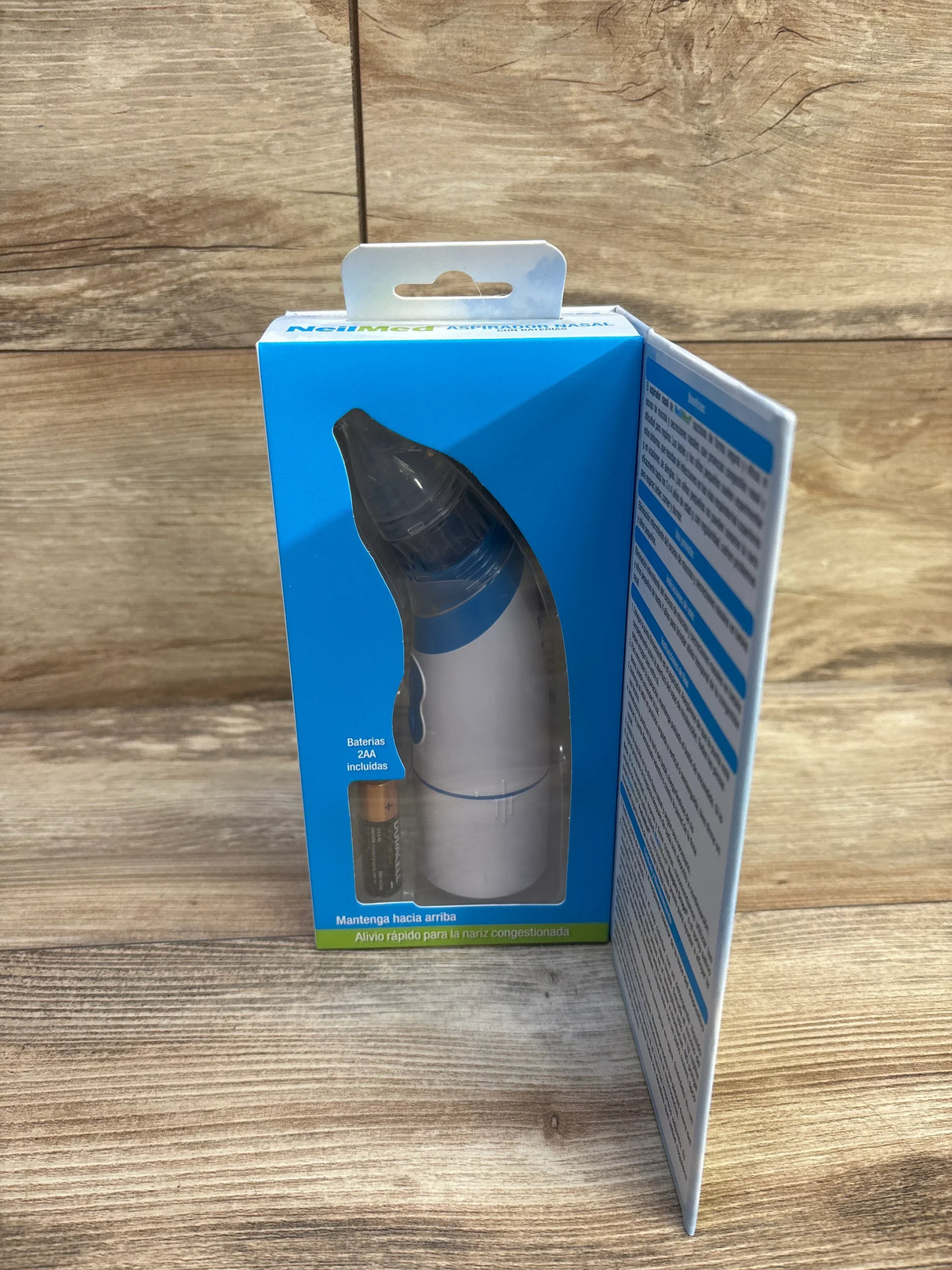 Angled front of NeilMed battery nasal aspirator in blue box showing clear tip, included AA battery, and open instruction flap