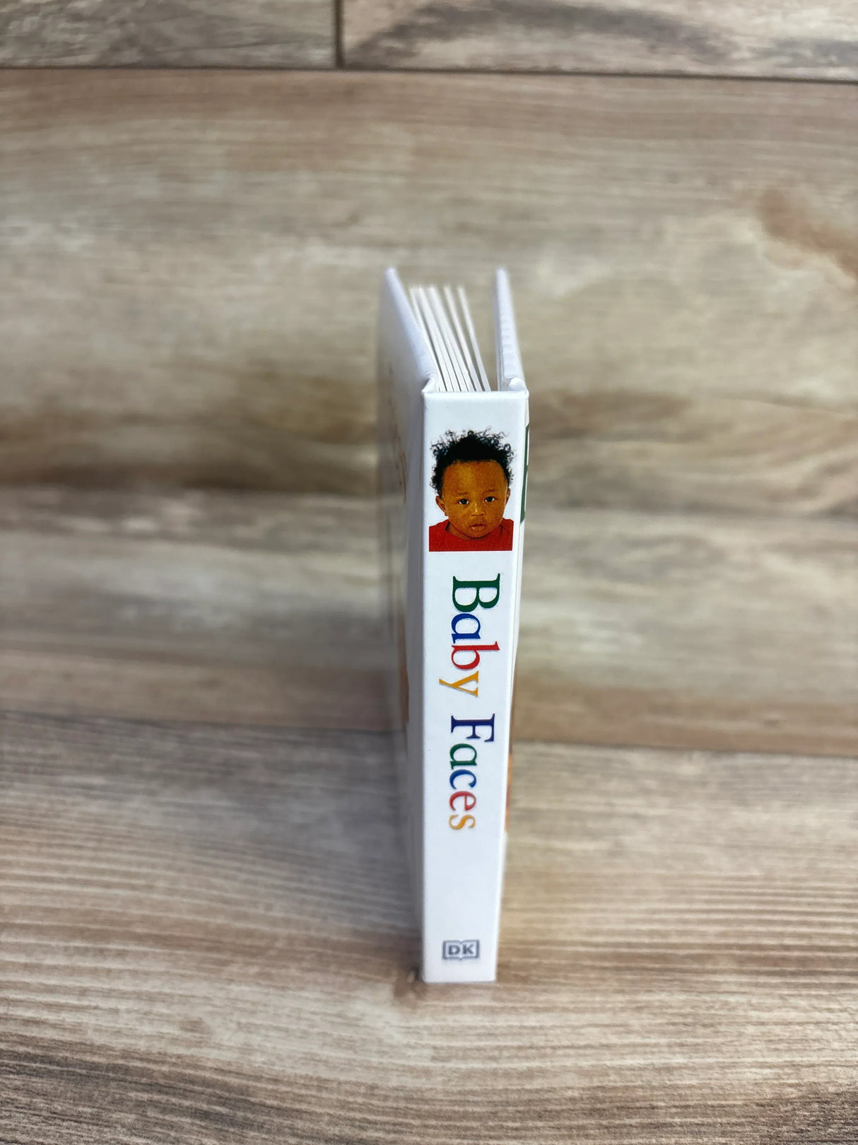 Spine view of Baby Faces DK board book showing colorful title text and infant cover photo standing on wood plank floor