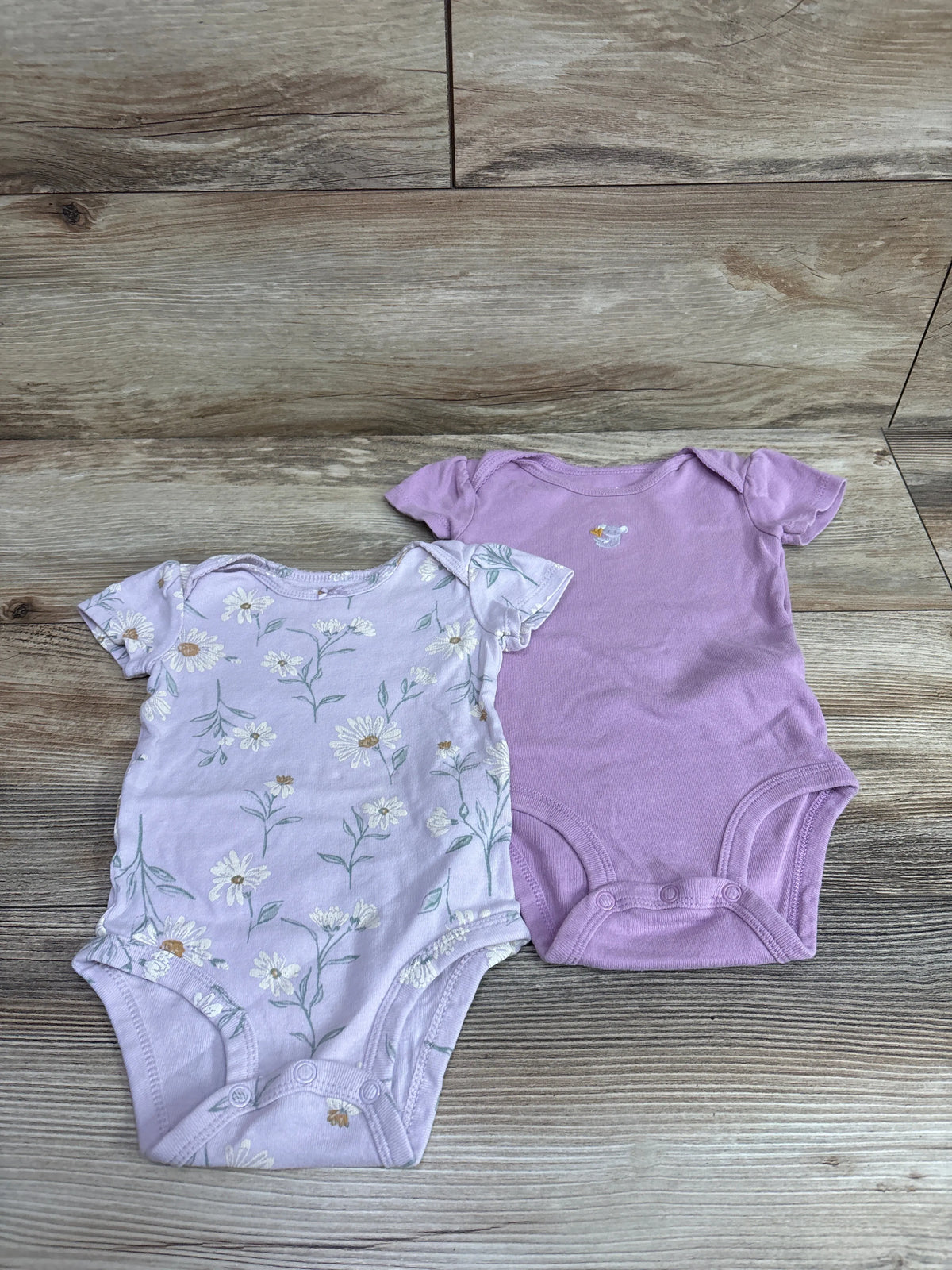 Two purple Carter's baby bodysuits, one solid and one with a floral print, on a wood background.