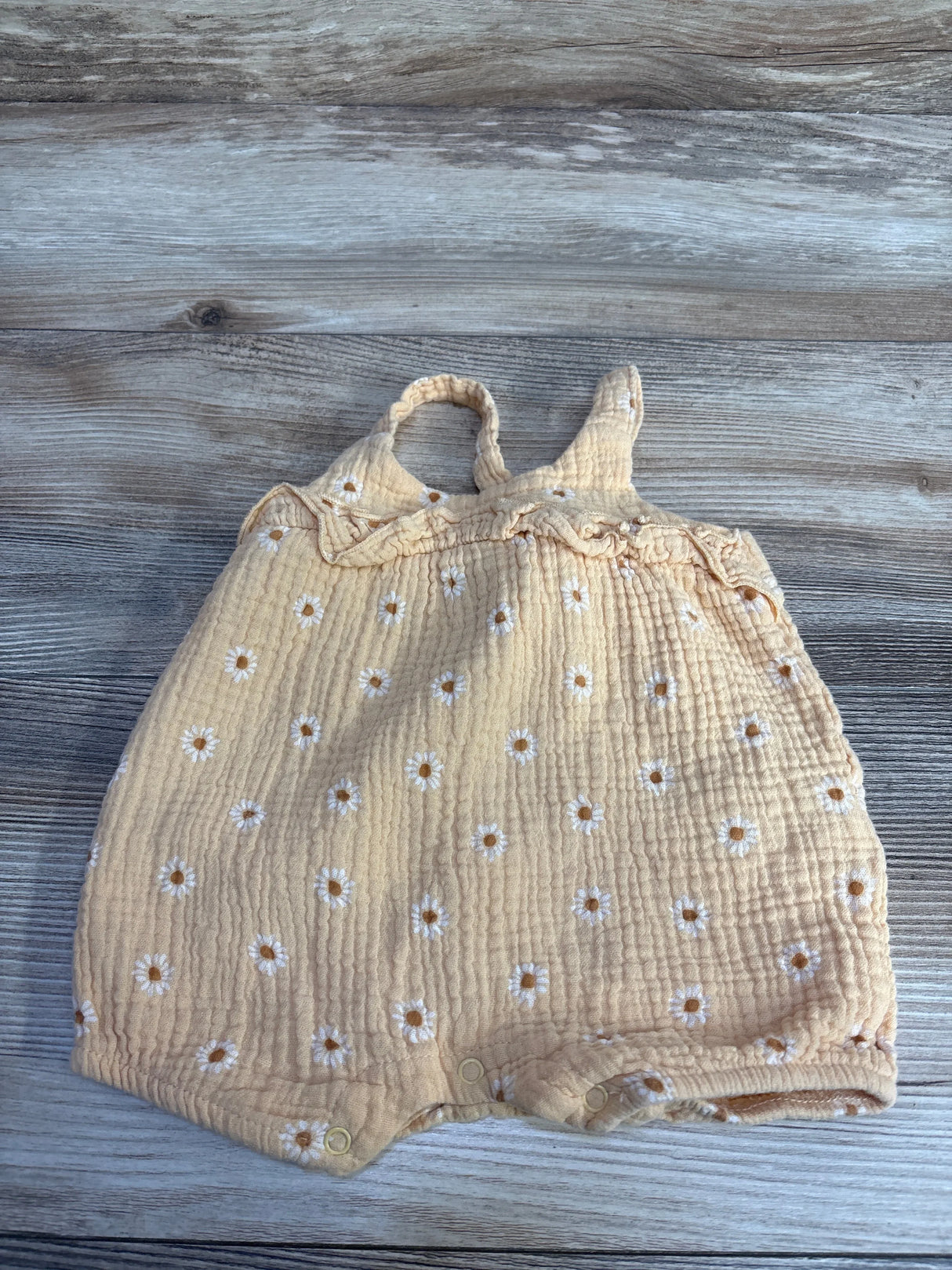 Flat lay of orange gauze baby romper 3 to 6 months with embroidered white daisies, ruffle straps and snap closures on wood