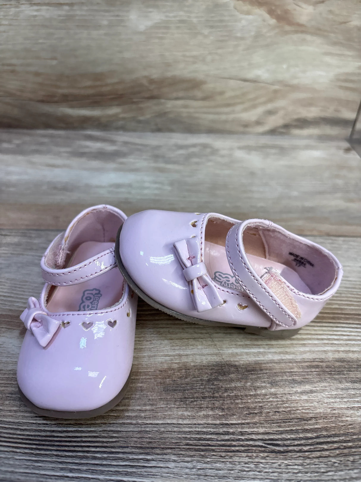 Three-quarter view of Teeny Toes pink patent Mary Jane flats showcasing heart-shaped cutouts, bows, and Velcro strap on a light wood background