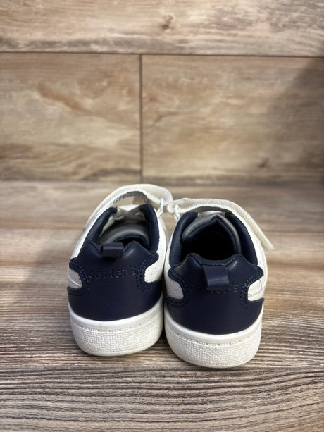 Rear view of Carter's Every Step Port toddler sneakers in white with navy heel panels and Velcro straps on wood-look background