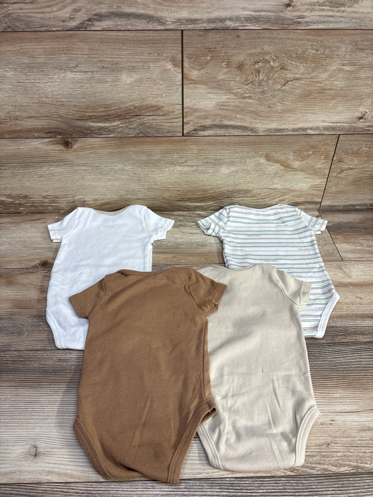 Four baby onesies in white, striped, beige, and brown laid out on wooden floor