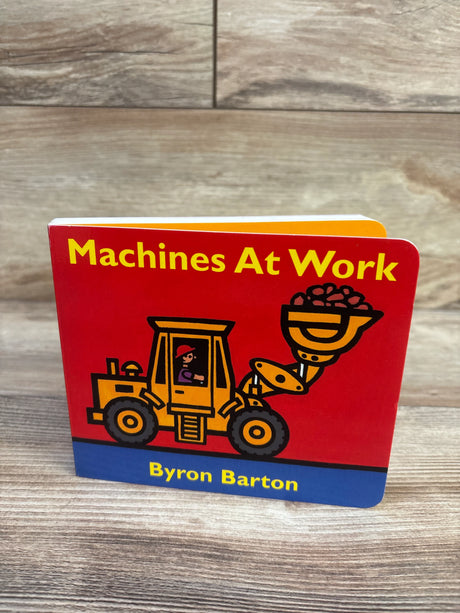 Front view of Machines At Work board book showcasing red cover with yellow loader illustration and author Byron Barton on wood background