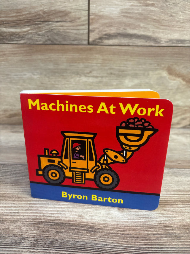 Front view of Machines At Work board book showcasing red cover with yellow loader illustration and author Byron Barton on wood background