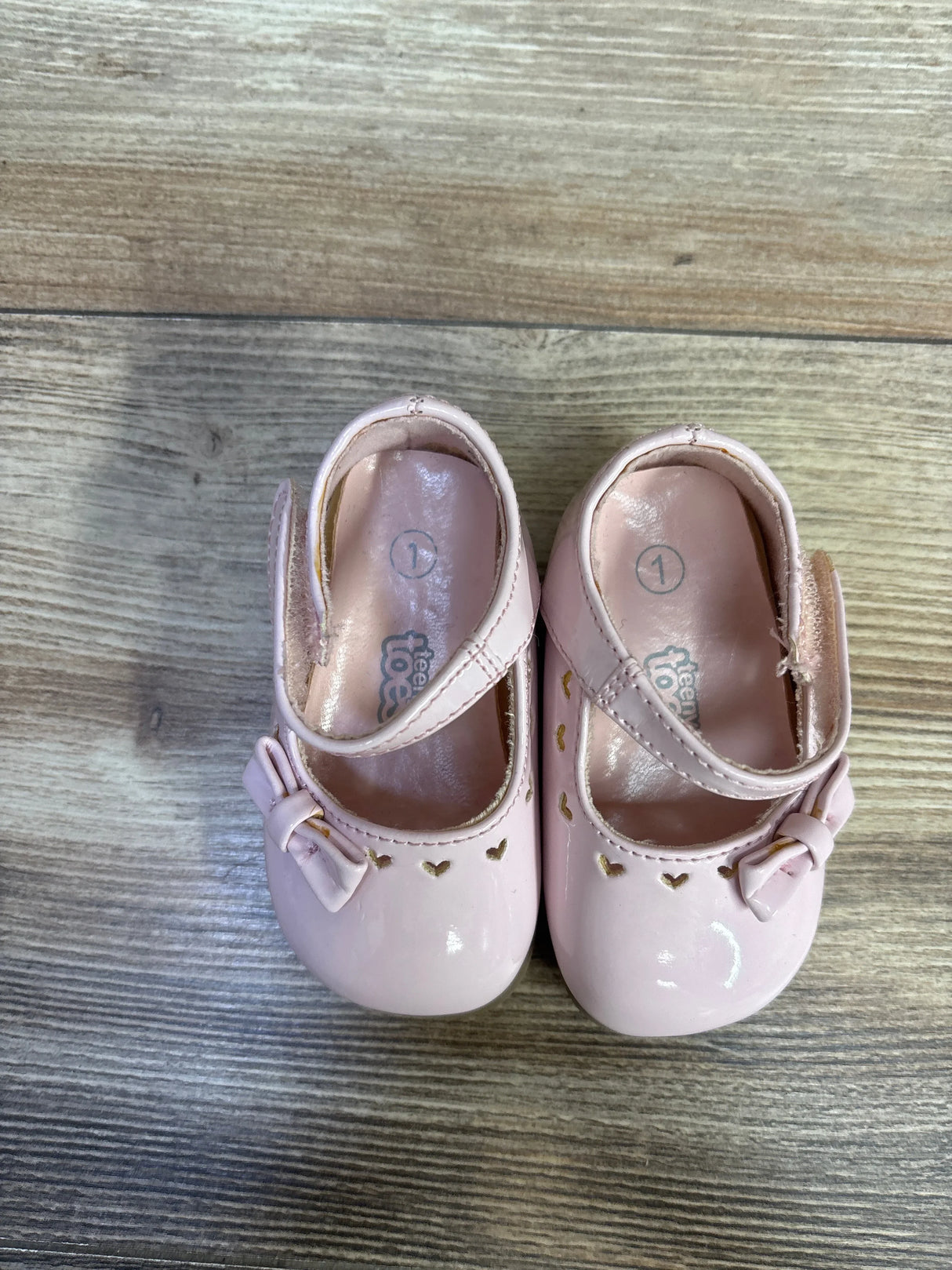Top view of pink patent Teeny Toes Mary Jane baby flats showing heart-shaped cutouts and bow accents on wood floor