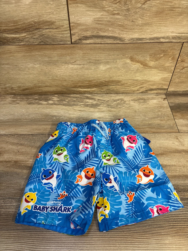 Flat lay of blue Baby Shark swim trunks showing elastic waistband and colorful shark characters on tropical-leaf print on wood floor