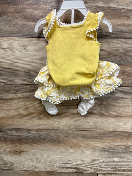 Yellow newborn 4-piece outfit with ribbed tank, pom-pom trim, tiered floral skirt, and white socks on wood background