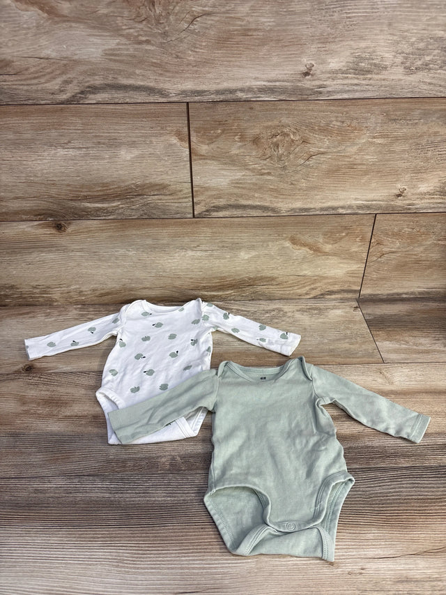 Flat lay of H&M organic 2-pack newborn bodysuits showing white apple-print and solid sage green on wood background