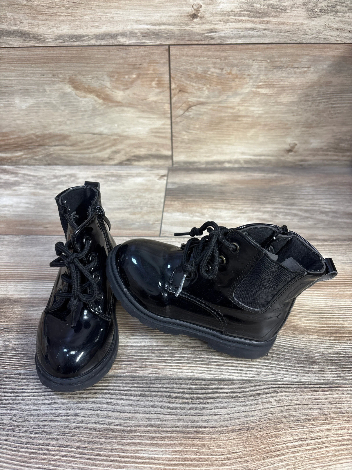 Side front view of kids' black faux leather lace up boots with glossy finish and chunky sole on wood background