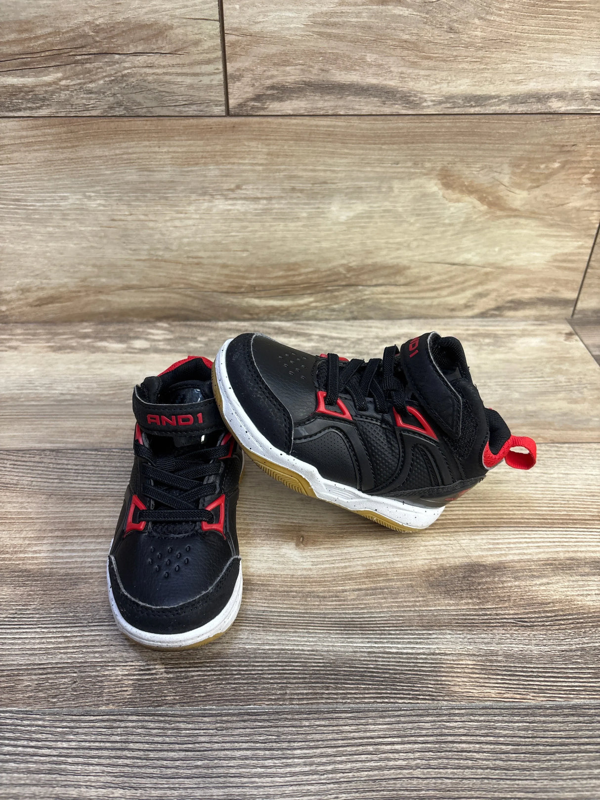 Front three quarter view of black AND1 high-top basketball shoes with red trim, velcro strap and gum sole on wood floor