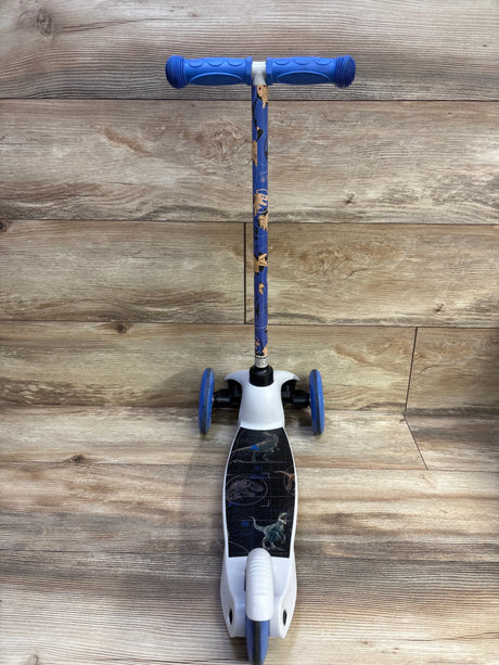 Front view of Jurassic World three-wheel tilt and turn scooter showcasing blue grips and dinosaur deck and stem graphics with wood-plank background