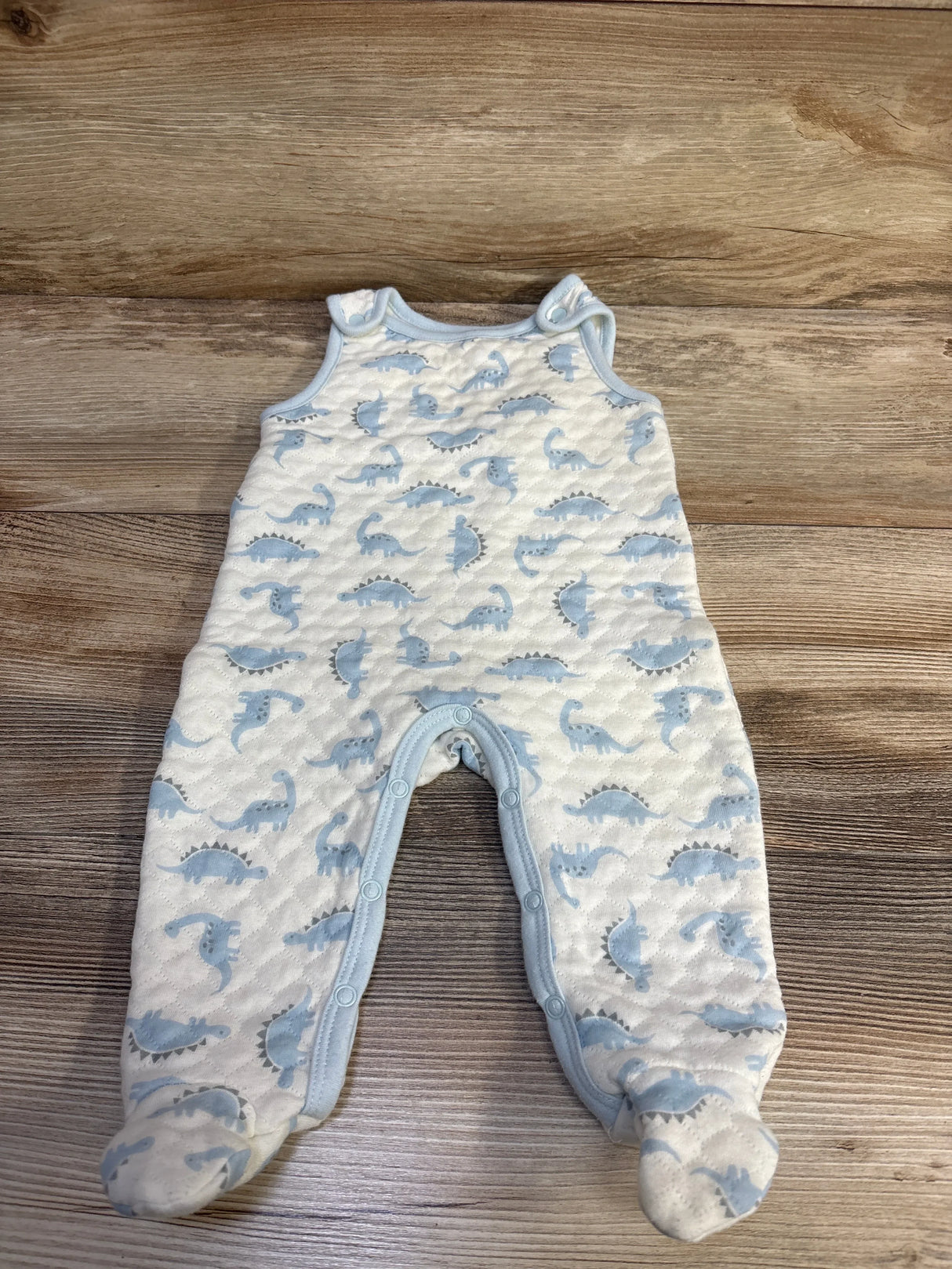 Flat lay of Minibean white footed jumpsuit with blue dinosaur print and snaps along legs on wood background
