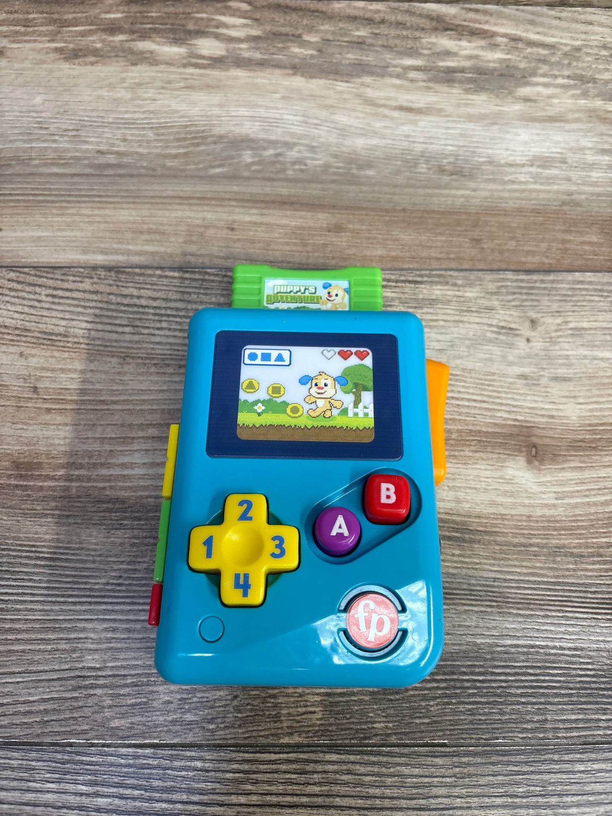 Top view of Fisher-Price Laugh and Learn Lil' Gamer with numbered d-pad, A and B buttons, and puppy game screen on wood