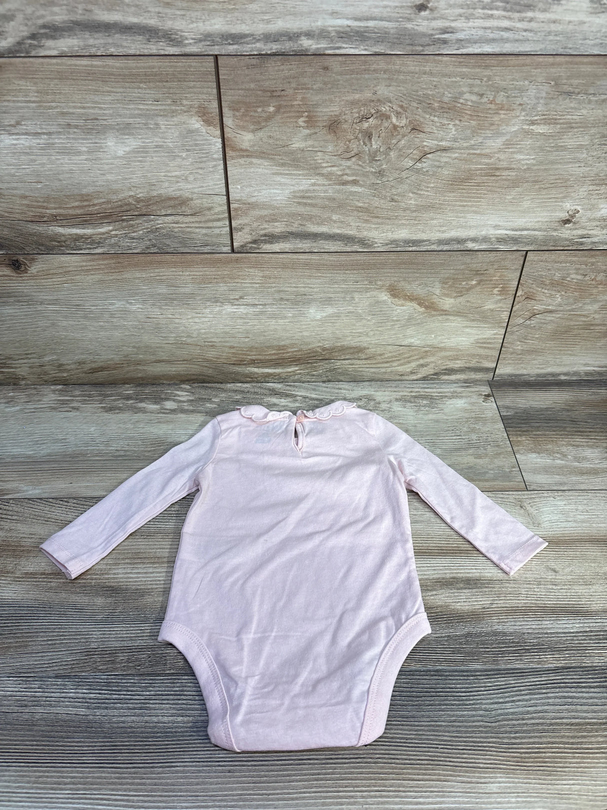 Flat lay of pale pink Oshkosh long-sleeve bodysuit showing scalloped Peter Pan collar and back keyhole on wood background