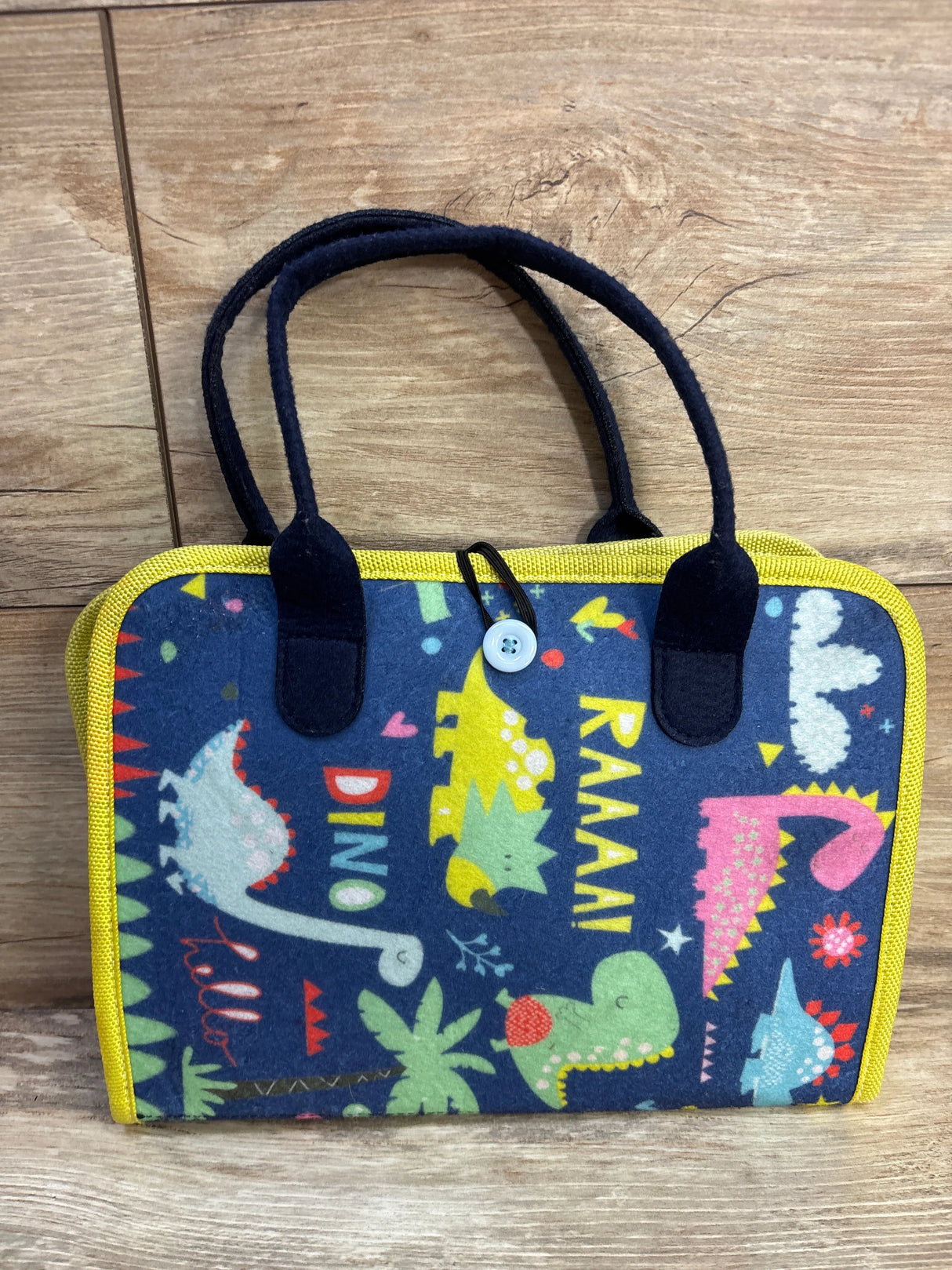 Front view of Hooku Montessori Busy Felt Book showing navy felt cover with colorful dinosaur prints, yellow trim, twin handles and button-loop closure on wooden floor