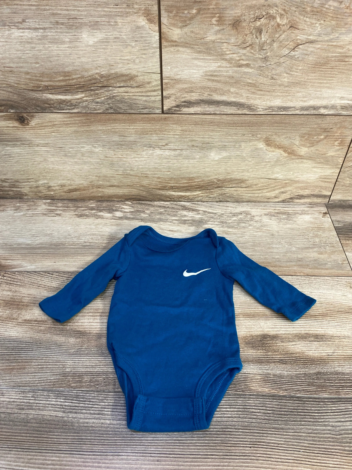 Flat lay of blue Nike long-sleeve baby bodysuit size 3M showcasing white swoosh on chest with wood floor background