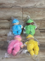 Overhead view of four Lakeshore cuddly washable dolls (blue, green, pink, yellow) in clear plastic bags on a wood plank surface