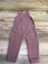 Front view of mauve knit overalls with buttoned straps, a front pocket, and ribbed cuffs on a wood background