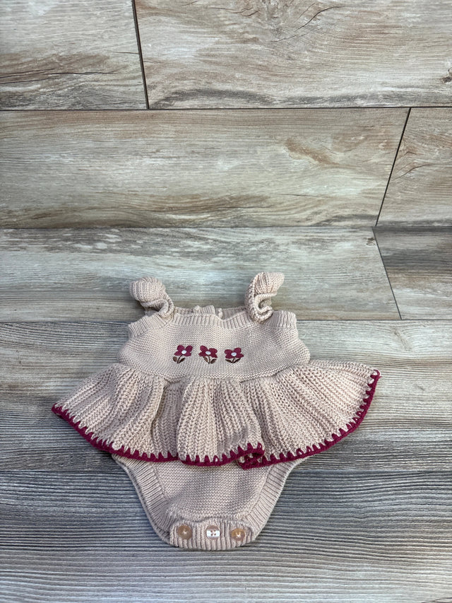 Flat lay of beige Nicole Miller knitted baby bodysuit showcasing embroidered flowers, ruffled skirt and maroon-trimmed hem on wood floor
