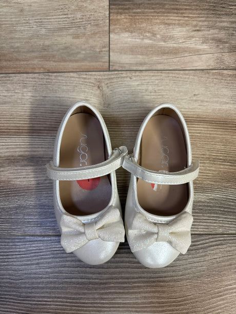 Top-down view of white toddler Mary Jane flats showcasing glitter bows and hook-and-loop ankle straps on wood floor