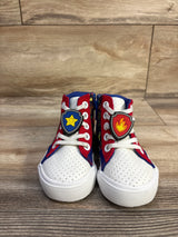 Front view of Paw Patrol high-top sneakers with white perforated toe, red-blue panels and shield lace charms on wood floor