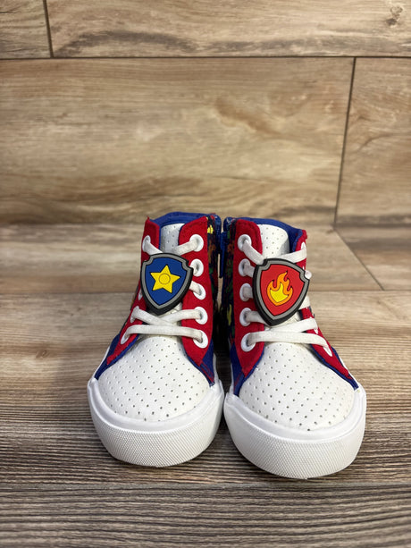 Front view of Paw Patrol high-top sneakers with white perforated toe, red-blue panels and shield lace charms on wood floor